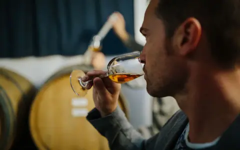 Mann verkostet bernsteinfarbenen italienischen Dessertwein in einem Glas, im Hintergrund Weinfässer.