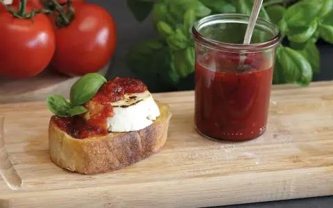 Bruschetta mit Ziegenkäse und Tomatenmarmelade, inspiriert von einem italienischen Tomatenmarmelade Rezept. Frische Tomaten und Basilikum im Hintergrund.