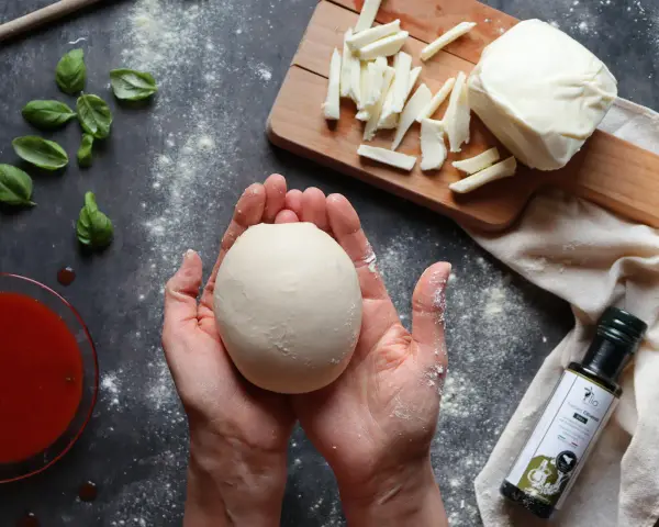 Hände formen Teig für ein Caputo Nuvola Rezept. Mozzarella, Basilikum und Tomatensauce liegen bereit.