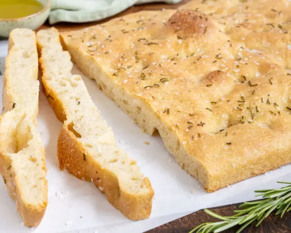 Frisch gebackene Focaccia, ein Klassiker unter den italienischen Brotsorten, mit grobem Salz und Rosmarin bestreut. Daneben liegen zwei geschnittene Stücke.