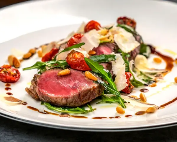 Ein saftiges Steak, italienisch zubereitet, mit Rucola, Kirschtomaten, Pinienkernen und Parmesanspänen auf einem weißen Teller.