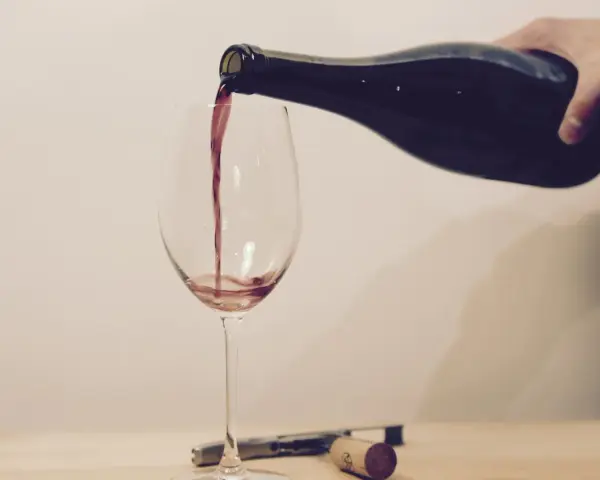 Ein Weinglas wird mit Rotwein gefüllt. Eine Hand hält die dunkle Flasche, während der Wein in einem dünnen Strahl ins Glas fließt. Ein Korkenzieher und ein Korken liegen daneben.