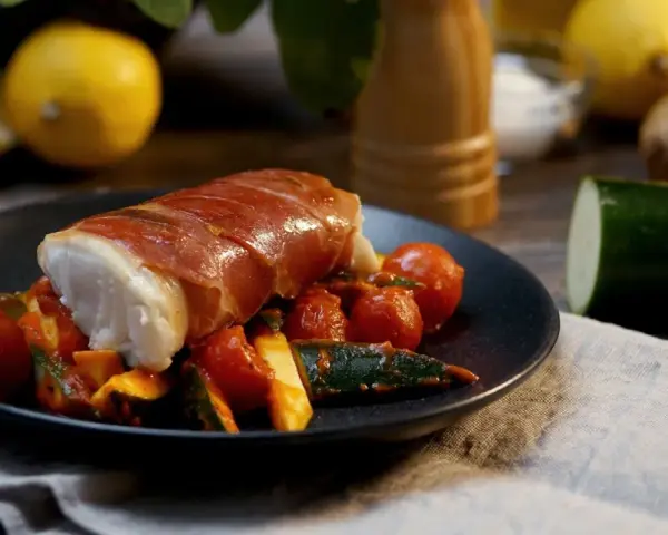 Kabeljau Saltimbocca: ein zartes Fischfilet, umwickelt mit luftgetrocknetem Schinken, serviert auf einem Bett aus Kirschtomaten und Zucchini.