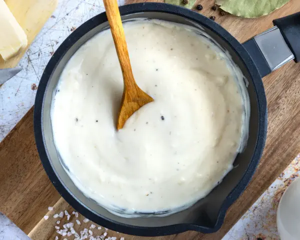 Cremige Béchamelsauce in einer Pfanne, bereit für Lasagne. Butter, Milch und Gewürze liegen daneben.