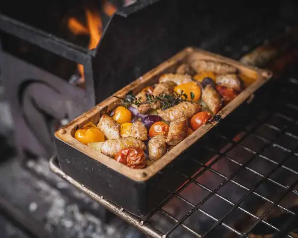 Salsiccia grillen in einer Keramikform mit Tomaten und Kräutern. Flammen lodern im Hintergrund.