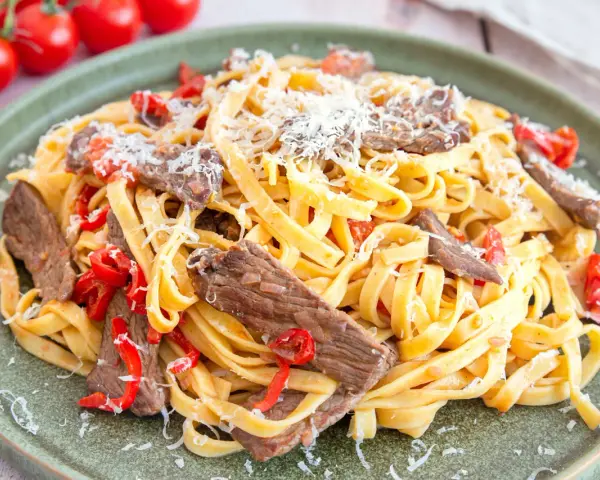 Pasta mit Rinderfiletstreifen, Tomaten und Parmesan auf einem grünen Teller.