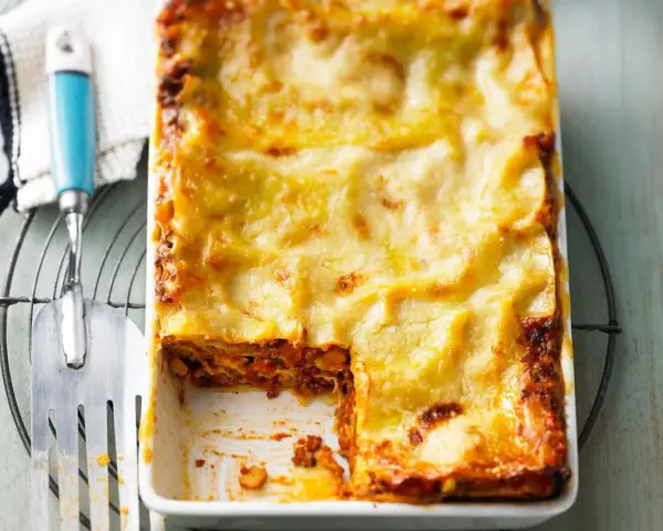 Eine frisch gebackene Lasagne al Ragu, goldbraun überbacken, wartet darauf, serviert zu werden. Ein Stück wurde bereits entnommen.