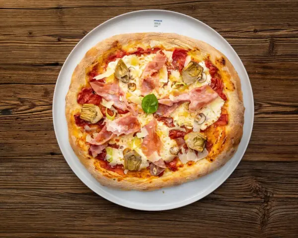 Eine köstliche Prosciutto Pizza mit Artischocken und Basilikum auf einem weißen Teller, serviert auf rustikalem Holz.