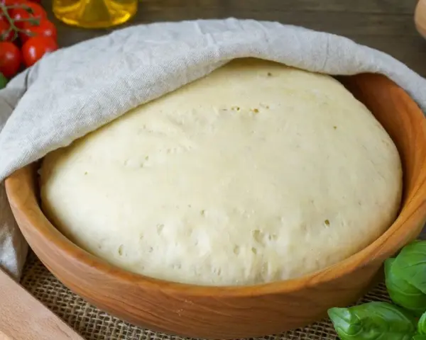 Frischer Pizzateig ruht in einer Holzschüssel, bedeckt mit einem Tuch. Tomaten und Basilikum im Hintergrund deuten auf die bevorstehende Zubereitung hin.