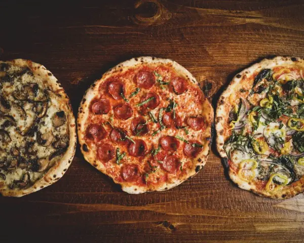 Drei italienische Pizzen: eine mit Pilzen, eine mit Salami und eine mit Gemüse, liegen auf einem Holztisch.