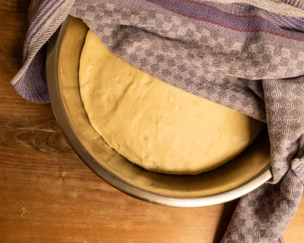 Pizzateig ruht in einer Schüssel, bedeckt mit einem gemusterten Tuch. Die Frage "wie lange pizzateig gehen lassen" beschäftigt den Bäcker.