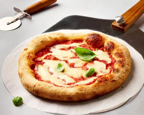 Frisch gebackene Pizza Margherita mit geschmolzenem Mozzarella und Basilikum. Daneben liegen ein Pizzaschneider und eine Pizzaschaufel, bereit, den perfekten pizzateig ausrollen.