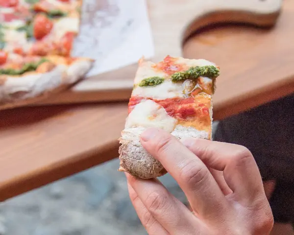 Eine Hand hält ein Stück Pizza mit Tomaten, Mozzarella und Pesto. So sieht eine köstliche Pizza aus!