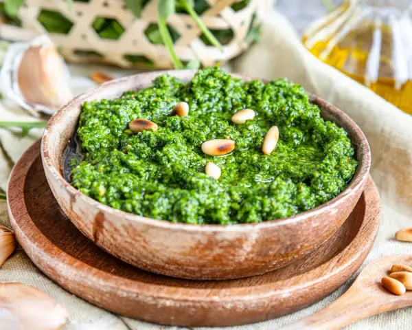 Frisches Rucola Pesto, garniert mit Pinienkernen, daneben Knoblauch und Olivenöl.