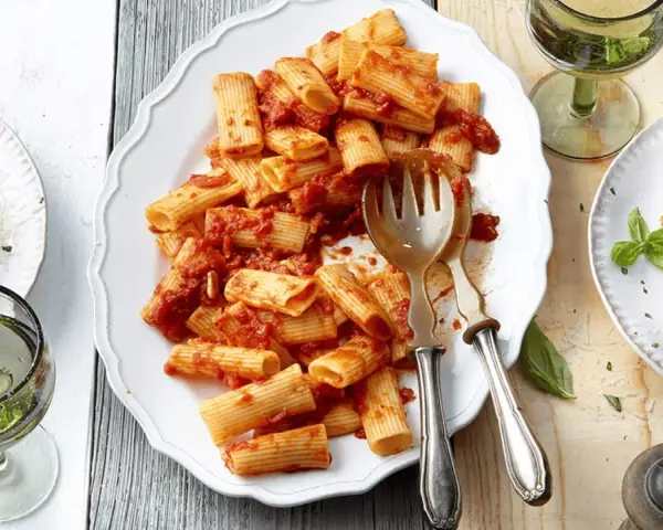 Ein Teller mit Rigatoni mit Tomatensauce, garniert mit Parmesan und Basilikum. Daneben ein Glas Weißwein.