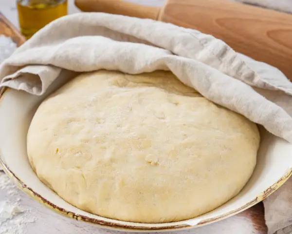 Frisch zubereiteter pizzateig mit trockenhefe ruht unter einem Tuch. Daneben liegen Salz und Olivenöl.