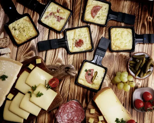 Raclette-Pfännchen mit geschmolzenem Käse, dazu Brot, Käse, Salami, Cornichons und Trauben. Perfekt für einen gemütlichen Abend mit **Wein zum Raclette**.