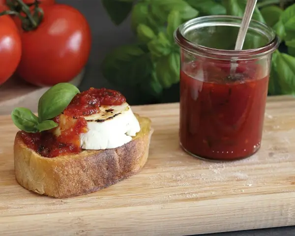 Bruschetta mit Ziegenkäse und Tomatenmarmelade, inspiriert von einem italienischen Tomatenmarmelade Rezept. Frische Tomaten und Basilikum im Hintergrund.