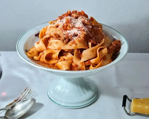 Ein Teller breiter Bandnudeln mit herzhaftem al ragu, bestreut mit Parmesan. Daneben ein Glas Rotwein und Besteck.