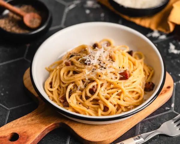 Eine Schüssel mit echter Carbonara, garniert mit geriebenem Parmesan und schwarzem Pfeffer. Daneben ein Holzlöffel mit Pfefferkörnern.