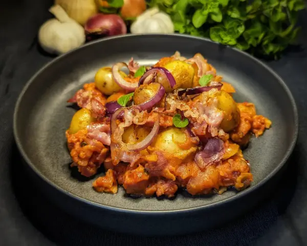 Herzhafte Salsiccia-Rezepte ohne Pasta: Kartoffeln, rote Zwiebeln und Speck in einer würzigen Tomatensauce, garniert mit frischen Kräutern.