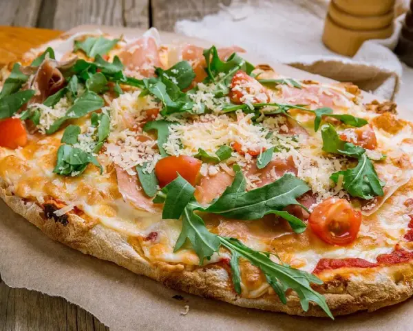 Eine köstliche Pinsa belegt mit Schinken, Rucola, Kirschtomaten und viel Käse auf rustikalem Holzuntergrund.