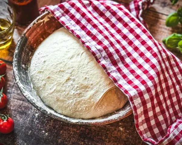 Frischer Pizzateig ruht in einer Schüssel, bedeckt mit einem rot-weiß karierten Tuch. Daneben liegen Kirschtomaten und ein Glas Olivenöl – die perfekten pizzateig zutaten.