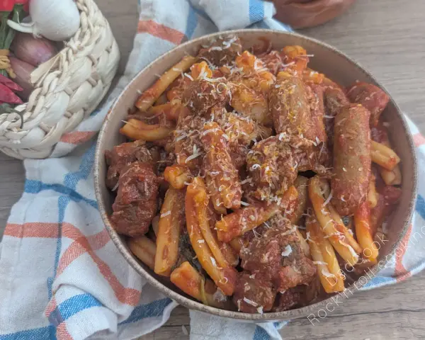 Herzhaftes Pasta Ragu mit Fleischstücken und geriebenem Käse, serviert in einer Schüssel auf einem karierten Tuch. Daneben ein Korb mit Knoblauch, Schalotten und Chilis.