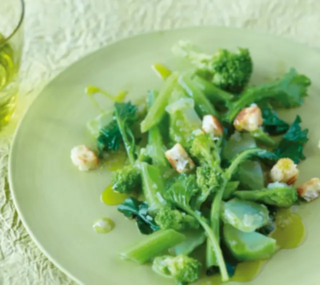 Ein Teller mit einem frischen Salat aus wildem Brokkoli, garniert mit Croutons und beträufelt mit Olivenöl. Daneben ein Glas grünes Getränk.