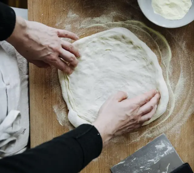 Hände formen einen Teig für ein Pizzateig Rezept auf einem Holzbrett. Mehl und ein Teigschaber liegen bereit.