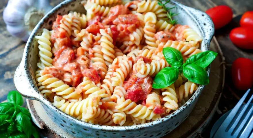 Fusilli-Nudeln in cremiger Tomaten-Sahne-Sauce, garniert mit frischem Basilikum. Daneben liegen Kirschtomaten und Knoblauch.
