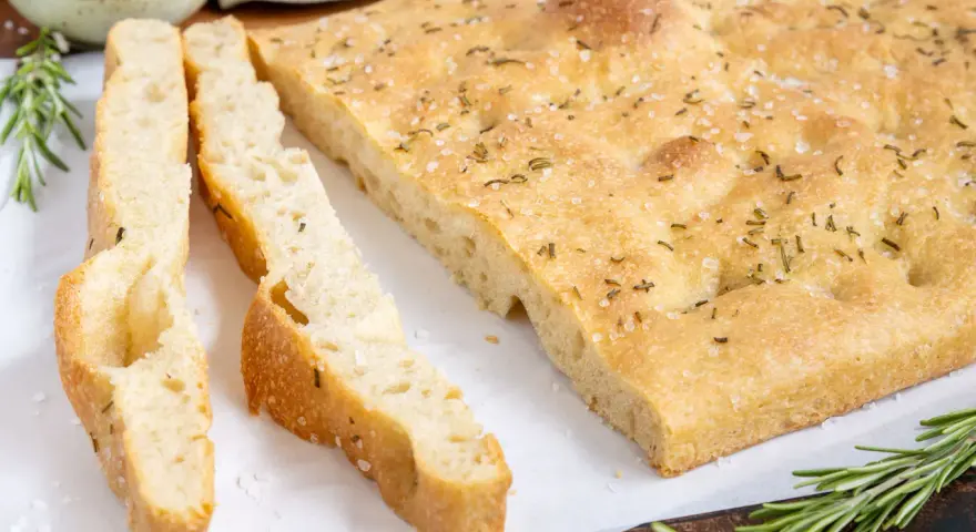 Frisch gebackene Focaccia, ein Klassiker unter den italienischen Brotsorten, mit grobem Salz und Rosmarin bestreut. Daneben liegen zwei geschnittene Stücke.