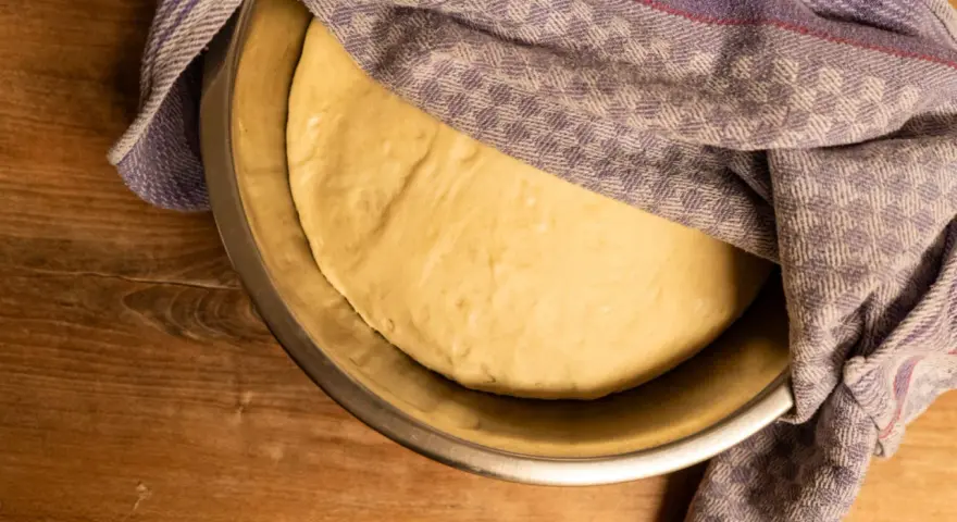 Frischer pizzateig für 4 Personen ruht in einer Schüssel unter einem karierten Tuch auf einem Holztisch.