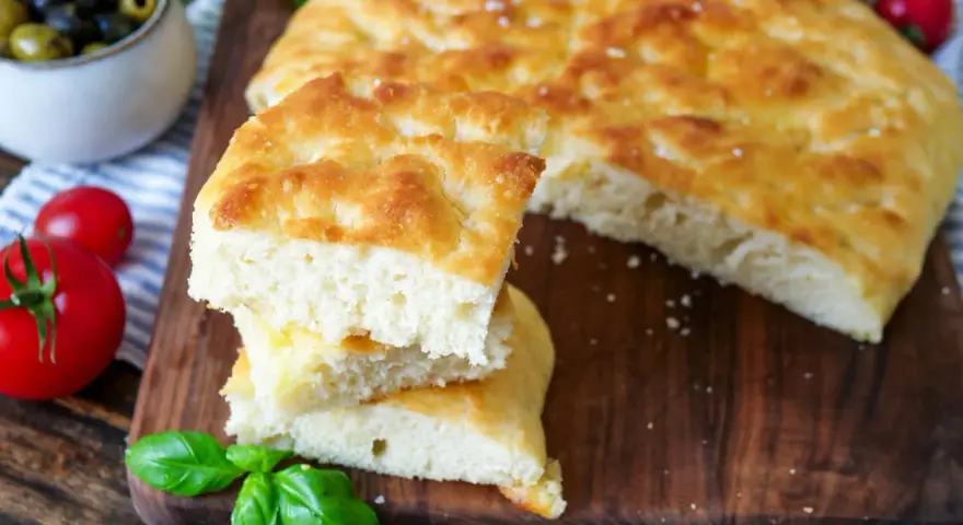 Frisch gebackener focaccia teig, aufgeschnitten und gestapelt, mit Tomaten und Oliven im Hintergrund.