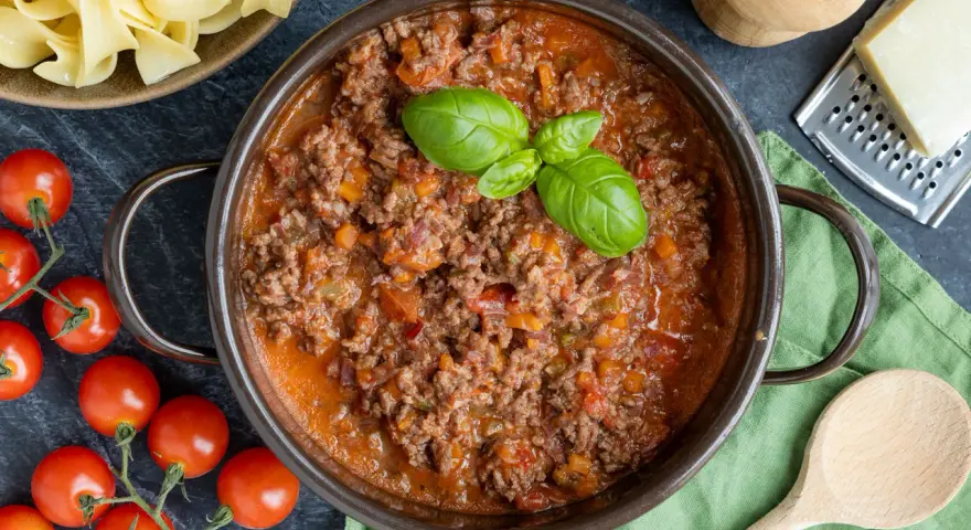 Herzhaftes ragù alla bolognese in einem Topf, garniert mit Basilikum. Daneben frische Tomaten, Bandnudeln und Parmesan.
