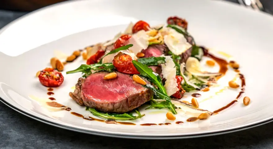 Ein saftiges Steak, italienisch zubereitet, mit Rucola, Kirschtomaten, Pinienkernen und Parmesanspänen auf einem weißen Teller.