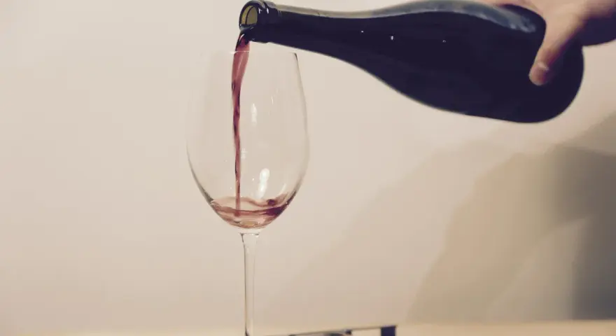 Ein Weinglas wird mit Rotwein gefüllt. Eine Hand hält die dunkle Flasche, während der Wein in einem dünnen Strahl ins Glas fließt. Ein Korkenzieher und ein Korken liegen daneben.