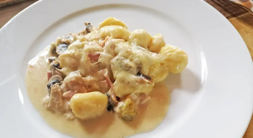 Gnocchi al forno mit cremiger Käsesauce, Pilzen und Schinken auf einem weißen Teller.