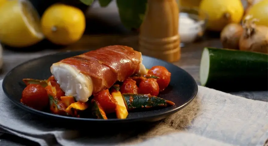 Kabeljau Saltimbocca: ein zartes Fischfilet, umwickelt mit luftgetrocknetem Schinken, serviert auf einem Bett aus Kirschtomaten und Zucchini.