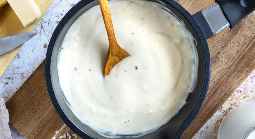 Cremige Béchamelsauce in einer Pfanne, bereit für Lasagne. Butter, Milch und Gewürze liegen daneben.