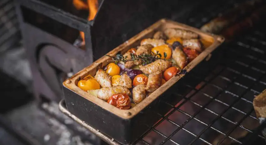 Salsiccia grillen in einer Keramikform mit Tomaten und Kräutern. Flammen lodern im Hintergrund.