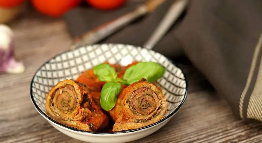 Zwei herzhafte involtini originalrezept in Tomatensoße, garniert mit Basilikum, in einer Schale auf Holztisch.