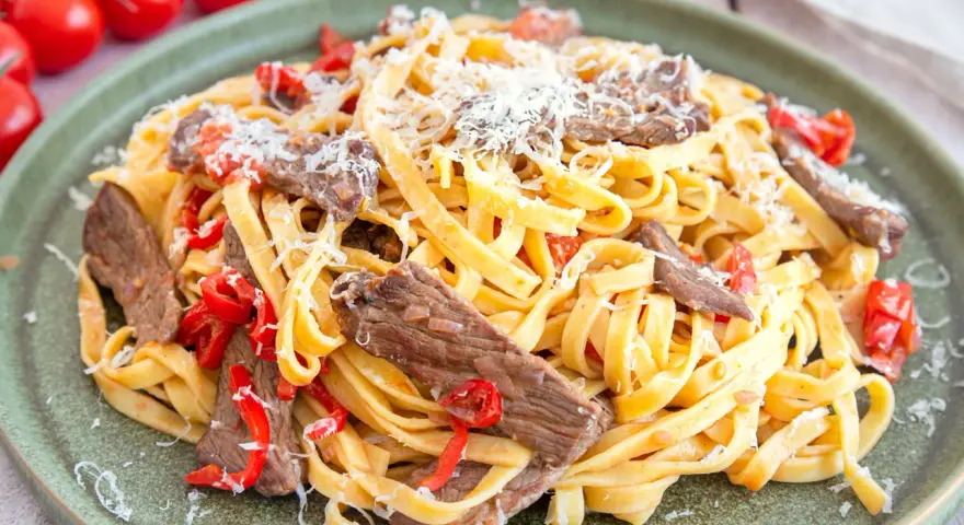 Pasta mit Rinderfiletstreifen, Tomaten und Parmesan auf einem grünen Teller.