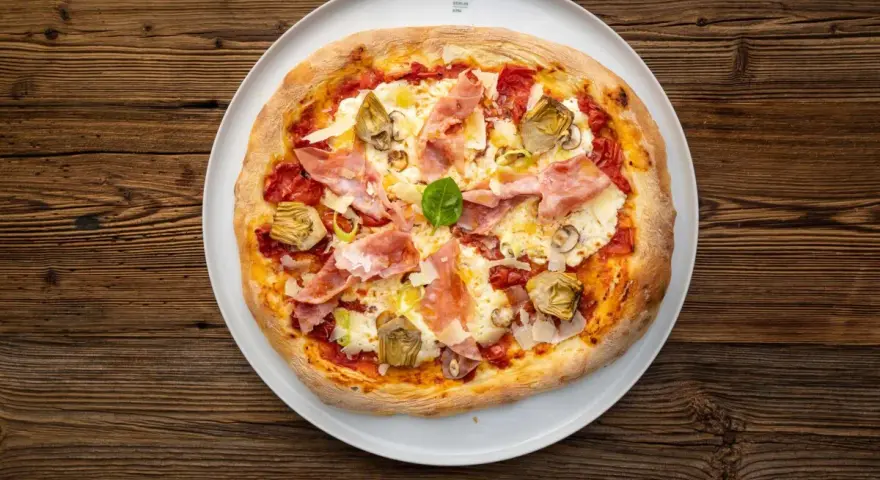 Eine köstliche Prosciutto Pizza mit Artischocken und Basilikum auf einem weißen Teller, serviert auf rustikalem Holz.
