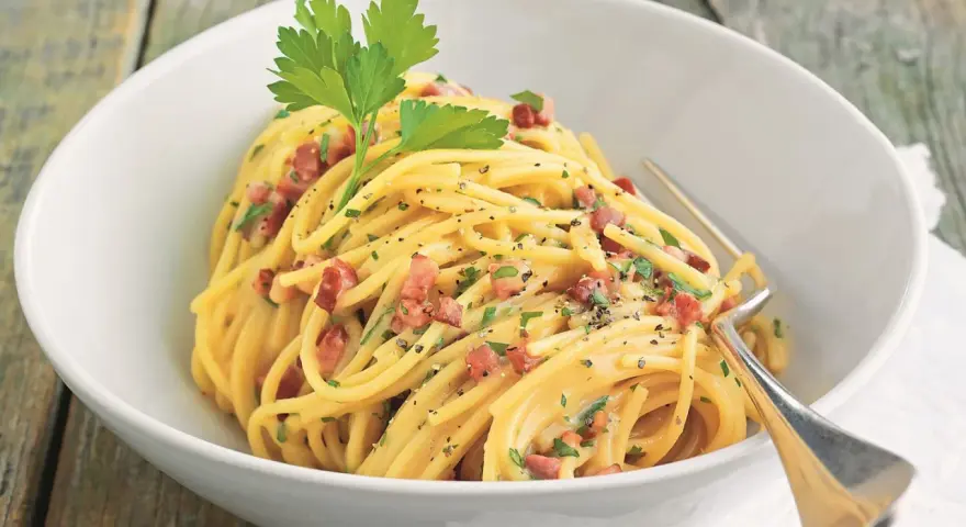 Spaghetti Carbonara mit knusprigem Schinken, garniert mit Petersilie und schwarzem Pfeffer, serviert in einer weißen Schüssel.