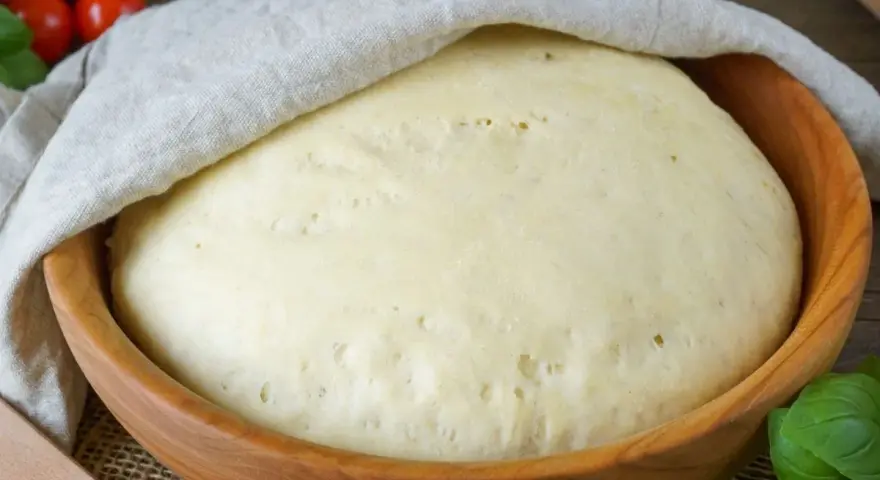 Frischer Pizzateig ruht in einer Holzschüssel, bedeckt mit einem Tuch. Tomaten und Basilikum im Hintergrund deuten auf die bevorstehende Zubereitung hin.