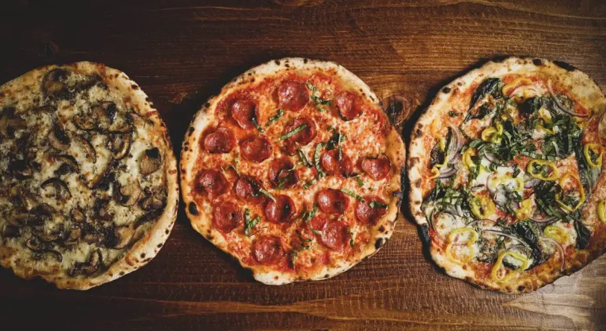 Drei italienische Pizzen: eine mit Pilzen, eine mit Salami und eine mit Gemüse, liegen auf einem Holztisch.