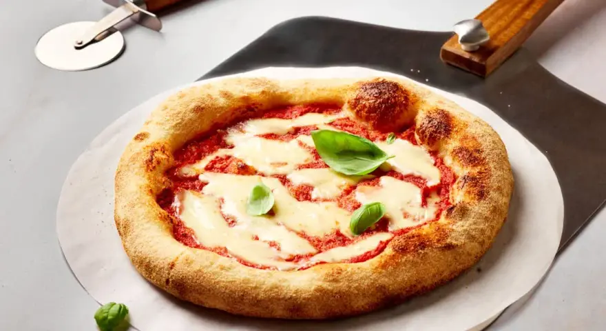 Frisch gebackene Pizza Margherita mit geschmolzenem Mozzarella und Basilikum. Daneben liegen ein Pizzaschneider und eine Pizzaschaufel, bereit, den perfekten pizzateig ausrollen.