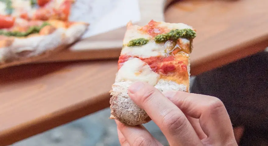 Eine Hand hält ein Stück Pizza mit Tomaten, Mozzarella und Pesto. So sieht eine köstliche Pizza aus!