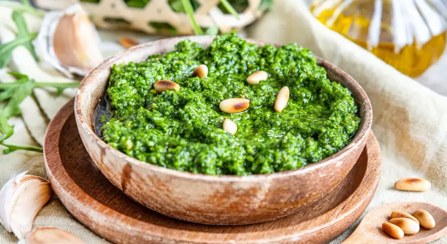 Frisches Rucola Pesto, garniert mit Pinienkernen, daneben Knoblauch und Olivenöl.